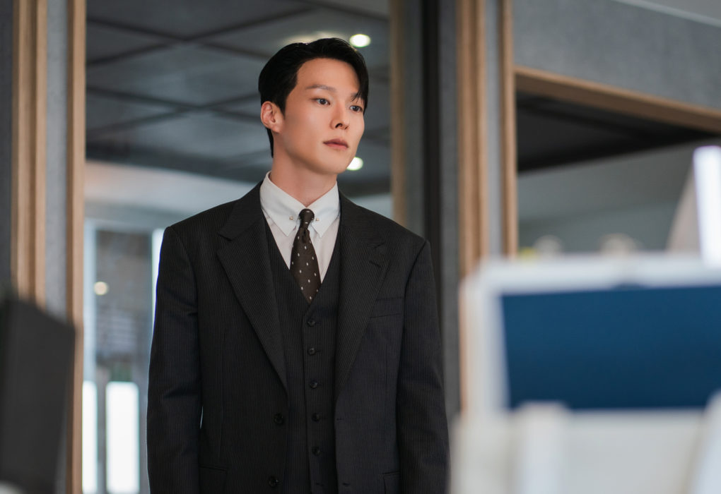 Jang Ki Yong, ator coreano é um Homem jovem veste terno escuro de três peças com camisa branca e gravata, em pé dentro de um escritório moderno. Ele olha para frente com expressão séria, transmitindo tensão e concentração, enquanto computadores aparecem desfocados em primeiro plano.