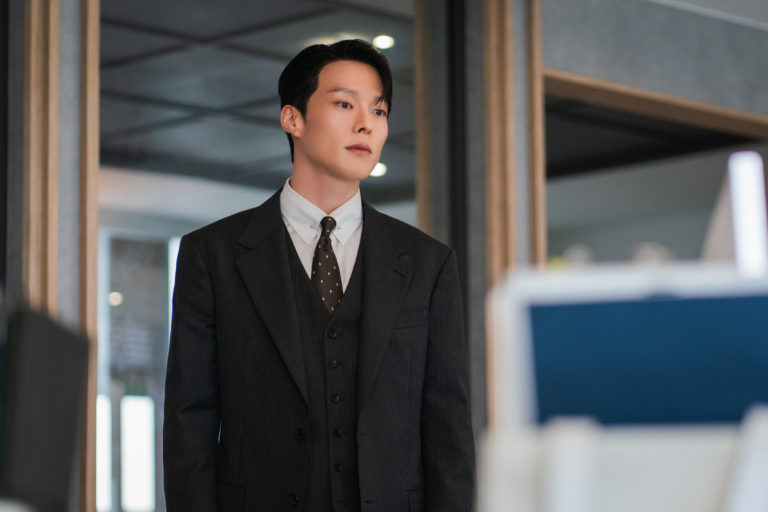 Jang Ki Yong, ator coreano é um Homem jovem veste terno escuro de três peças com camisa branca e gravata, em pé dentro de um escritório moderno. Ele olha para frente com expressão séria, transmitindo tensão e concentração, enquanto computadores aparecem desfocados em primeiro plano.