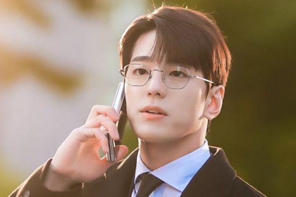 Kim Min Gue Homem de óculos e terno preto fala ao celular em uma cena ao ar livre, iluminada pela luz do sol.