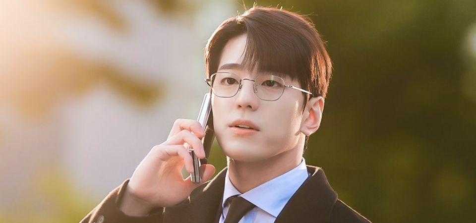 Kim Min Gue Homem de óculos e terno preto fala ao celular em uma cena ao ar livre, iluminada pela luz do sol.
