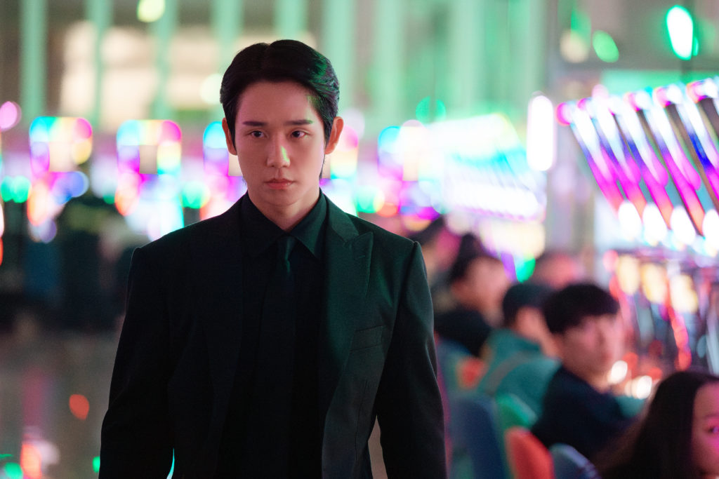 Jung Hae-in, caracterizado como Jang Tae-ha, aparece sério e vestido com terno preto em uma casa de jogos iluminada por luzes coloridas em cena do K-drama Um Grude de Amor, da Netflix.