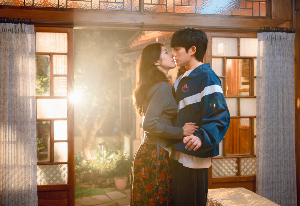 O ator coreano Jung Hae-in e a atriz coreana Ha Young se aproximam para um beijo em cena romântica do K-drama Um Grude de Amor, da Netflix, dentro de uma casa iluminada pela luz do sol.