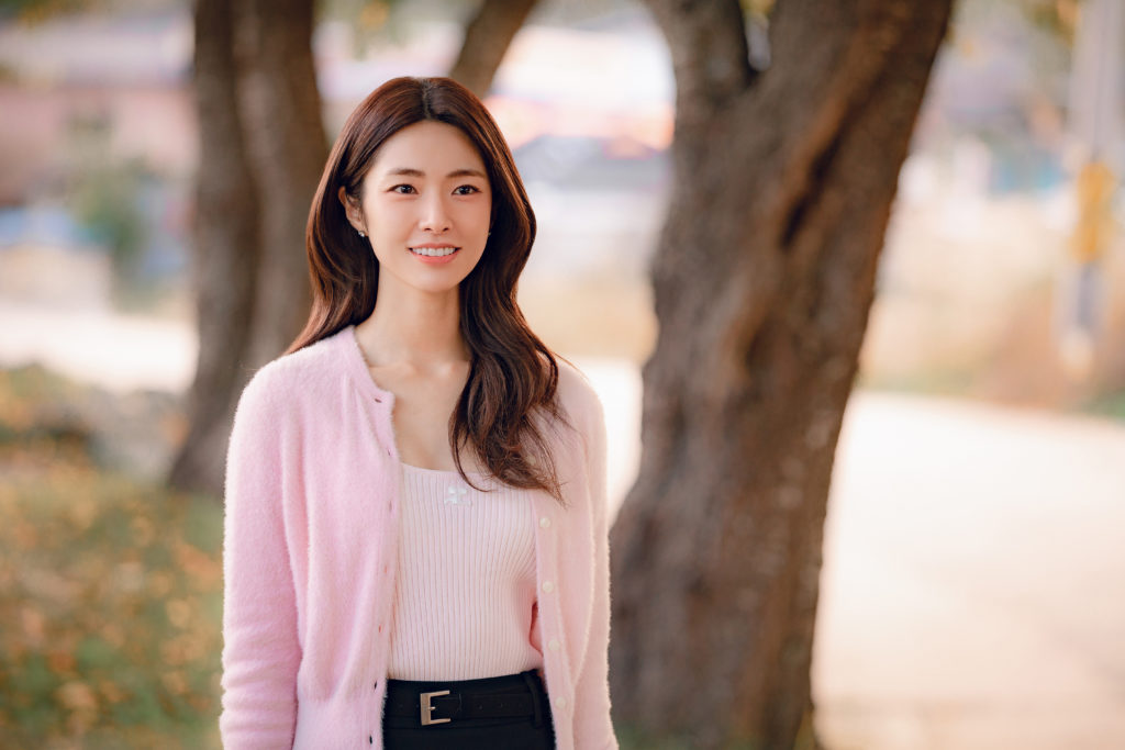 Ha Young, caracterizada como Go Eun-sae, sorri enquanto veste um cardigã rosa em uma cena ao ar livre do K-drama Um Grude de Amor, da Netflix.